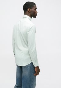 Šviesiai žalia ilgomis rankovėmis marškiniai su struktūruota apykakle, lieso silueto ir su sagų detalėmis; derinami su laisvai krentančiomis mėlynomis džinsinėmis kelnėmis.