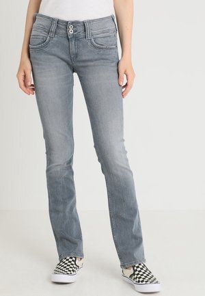 Persona con jeans ajustados gris desteñido y zapatos slip-on a cuadros en blanco y negro, de pie frente a un fondo claro y liso.