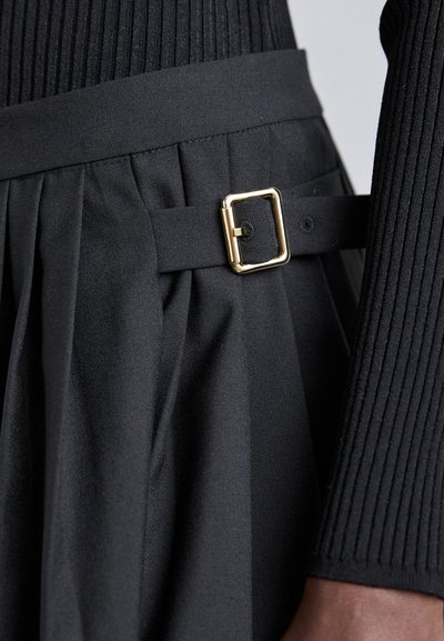 Schwarz gekleidetes Kleid mit einem gefältelten Rock und einem breiten Bund, verziert mit einer Metallschnalle und einer glatten Textur. Vertikale Rippenschichtung.
