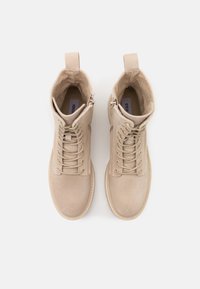 Beige ruskind ankelstøvler med rund tå, der har snørebånd, en sidelås og en løkke på hælen. Glat tekstur og syede detaljer.