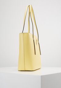 Żółta torba na ramię ze skóry, z długimi paskami, kwadratowym kształtem i gładką teksturą. Na froncie znajduje się okrągłe logo oraz kontrastowe przeszycia.
