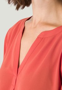 Blouse corail avec un col en V et des manches courtes, fabriquée en tissu léger au toucher doux et avec des détails de couture minimaux.