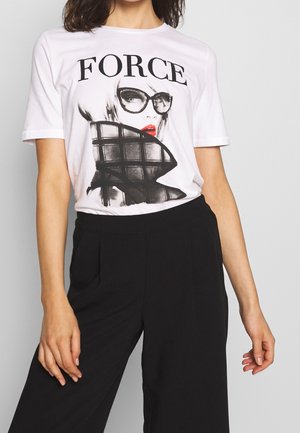 Woman wearing a white T-shirt with a graphic of a woman in glasses and red lipstick under the word "FORCE," paired with black pants.