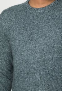 Gros plan sur une personne portant un pull en tricot doux et gris avec un col rond, mettant en valeur la texture et les détails du tissu.