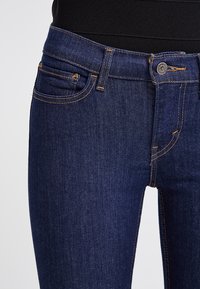 Jeans de mezclilla azul oscuro con un diseño ajustado, que presentan costuras naranjas, un botón en la parte frontal y cinco bolsillos. Textura suave y corte clásico.