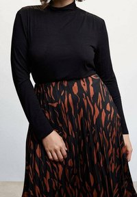 Mujer vestida con una blusa negra de manga larga metida dentro de una falda plisada negra con estampado abstracto de animales en naranja, de pie contra un fondo liso.