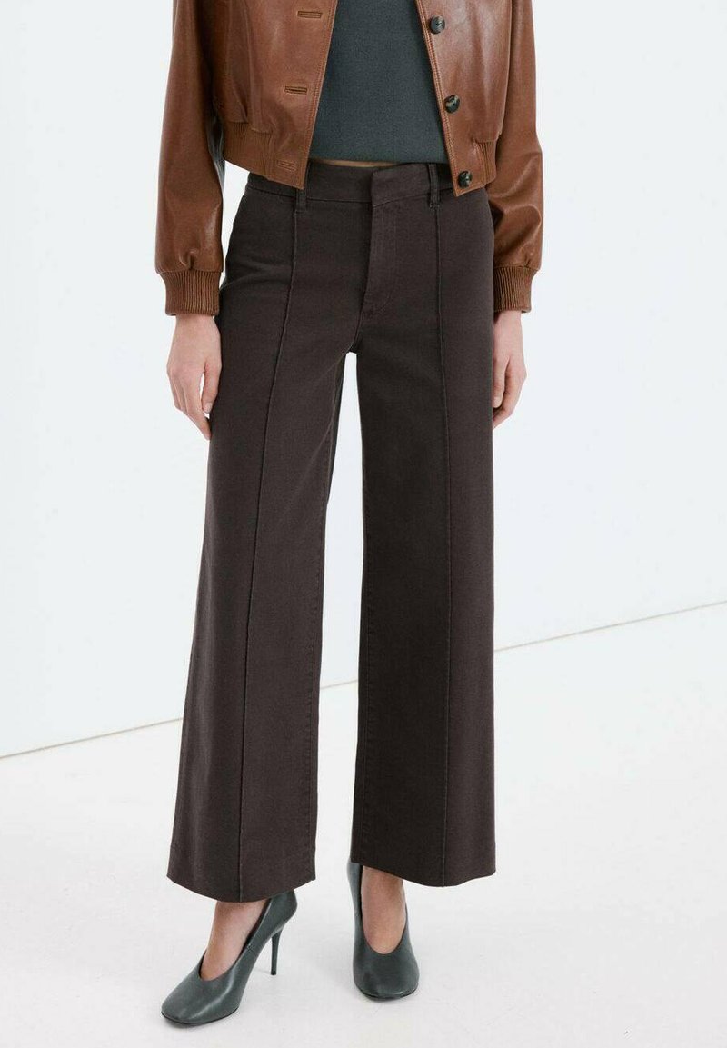 Pantaloni di jeans marroni a gamba larga con cuciture frontali, abbinati a un top grigio aderente e una giacca di pelle marrone, completati da décolleté verdi.