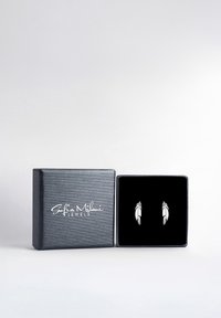 Silverfärgade fjäderformade örhängen visade i en låda klädd med svart sammet, med en strukturerad mörkgrå utsida och en vit logotyp.