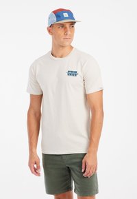 Bežinis crewneck marškinėliai su mėlynu grafiniu užrašu "PRO TEST." Derinami su alyvuogių žaliais šortais. Asmuo dėvi daugiaspalvę kepurę.