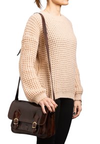 Satchel in pelle marrone con due fibbie, tracolla e finitura lucida. Maglione in maglia waffle di colore crema, pantaloni neri aderenti.