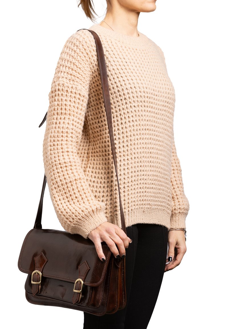 Satchel in pelle marrone con due fibbie, tracolla e finitura lucida. Maglione in maglia waffle di colore crema, pantaloni neri aderenti.