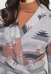 Cardigan envolvente en gris, con un patrón geométrico en tonalidades de rosa, negro y verde. Incluye un cinturón de lazo y una textura suave.