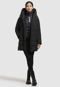 Cappotto lungo imbottito nero con un ampio cappuccio, dotato di chiusura con zip, e una giacca interna aderente. Indossato sopra leggings neri e stivali.