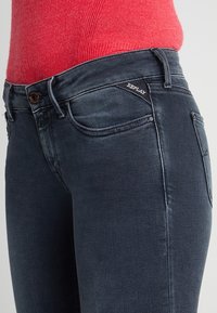 Jeans de mezclilla oscura con un diseño ajustado, que presentan un lavado sutil, un pequeño logo en el bolsillo trasero y herrajes metálicos clásicos en los botones.