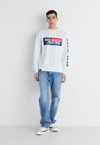Sudadera blanca con un estampado gráfico en rojo y azul que dice "RED BULL RACING". Combinada con jeans de color azul claro y zapatillas blancas.