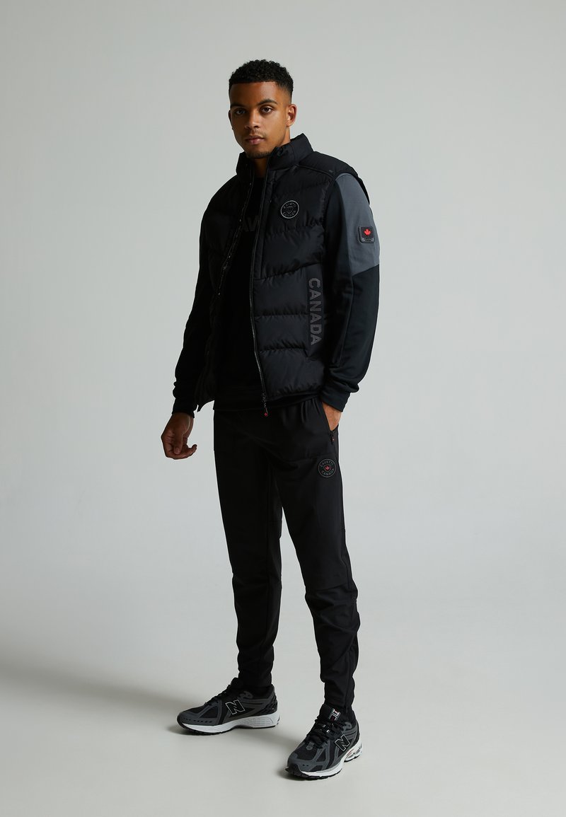 Doudoune noire avec accents gris, texte "CANADA" et logo. Pantalons noirs avec patch circulaire. Chaussures de sport noires avec détails gris.