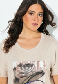 Camiseta beige de manga corta con un estampado gráfico de un sombrero de paja con acentos azules. Textura suave y ajuste relajado.