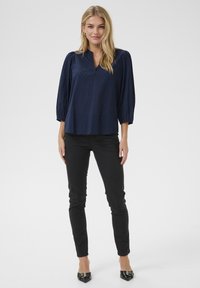 Blusa blu navy con scollatura a V, motivo strutturato e maniche a palloncino, abbinata a jeans neri slim-fit e décolleté nere con punta. SAP