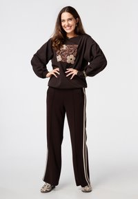 Un sweat-shirt marron avec broderies florales et manches ballon, associé à un pantalon marron avec des rayures blanches sur le côté, et des chaussures à imprimé léopard.