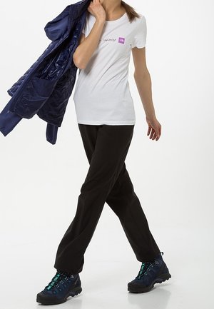 White t-shirt with purple logo, black pants, and navy puffer jacket. Dark hiking shoes with turquoise accents. Model walking.