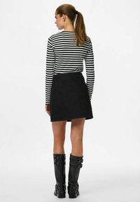 Femme avec queue de cheval portant un haut à manches longues rayé noir et blanc, une jupe noire et des bottes noires montantes avec boucles, debout de dos.