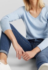 Vaaleansininen v-kaula-aukollinen neule 3/4-hihasilla, pehmeä tekstuuri, yhdistetty tummansinisiin farkkuihin, istuma-asennossa, valkoiset tennarit näkyvissä.