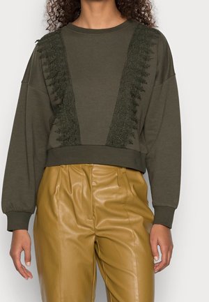 Kvinde iført olivengrøn langærmet sweatshirt med blonder og højtaljede læderbukser i sennepsgul, stående med afslappede hænder.