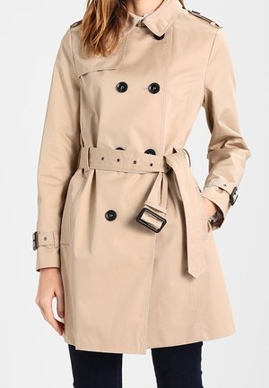 Vrouw draagt een beige dubbelgeknoopte trenchcoat met zwarte knopen en een ceintuur in de taille, staand met één hand in de zak.