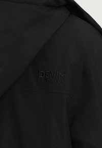 Czarny materiał z bliska, na którym widać tłoczone logo "DENIM TOM TAILOR" przeszyte na odzieży, z widocznym szwem.