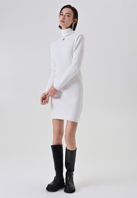 Donna che indossa un vestito a maglia bianco con collo alto, stivali neri fino al ginocchio, orecchini a cerchio dorati e una collana con pendente dorato, in piedi con le mani unite.