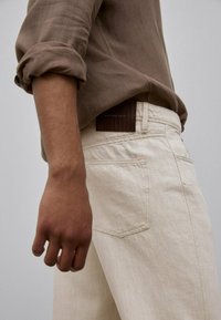 Beige farkut, joissa on sileä pinta, takatasku ja tumma ruskea merkkileather-tagi vyötäröllä. Vaaleanruskea pitkähihainen paita.