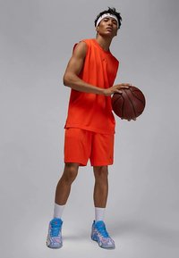 Oranje mouwloos sporttopje en bijpassende korte broek met zwart logo, houdt een basketbal vast. Draagt sneakers met roze en blauwe accenten.