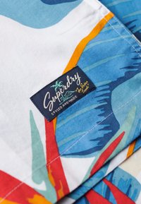 Bomuldsstof med et farverigt blomsterdesign, som indeholder blå, røde og grønne nuancer. Inkluderer et mærketikette "Superdry" på en navy baggrund.