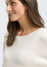 Pullover beige chiaro con scollo rotondo, caratterizzato da un motivo testurizzato e una lucentezza sottile. Il tessuto morbido si adagia delicatamente sulle spalle.