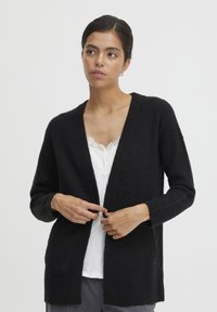 Svart cardigan med en lös passform, öppen framtill, långa ärmar, ribbad mudd och textilstruktur. Bärs över en vit spetsblus.