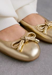 Ballerine dorate con punta rotonda e un dettaglio decorativo a fiocco con design annodato e accenti metallici. Finitura liscia e texturizzata.