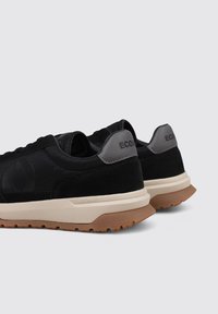 Zapatillas deportivas negras con una parte superior de ante y textil, un acento en el talón gris y una suela de goma texturizada en color marrón.