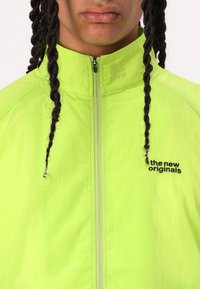 Neon green zip-up jacket with textured fabric; features “the new originals” logo in black on the left chest and a high collar.
