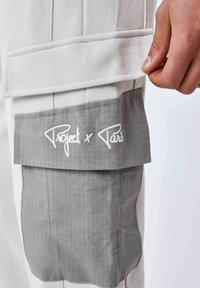 Witte, gestructureerde broek met een grijze stoffen zak en witte geborduurde "Project x Paris" branding aan de zijkant.