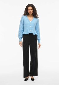 Hellblauer, kurz geschnittener Cardigan aus weichem, strukturiertem Material mit V-Ausschnitt und drei Knöpfen, kombiniert mit schwarzen weit geschnittenen Hosen und Absatzschuhen.
