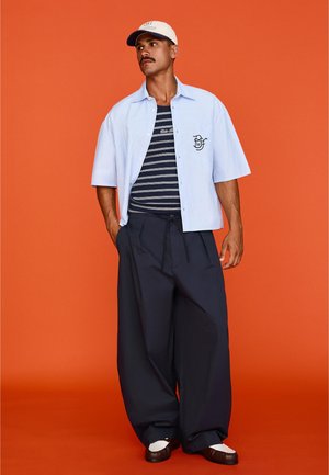 Mies, jolla on valkoinen lippalakki, vaaleansininen lyhythihainen nappipaita tummansinisen raitaisen t-paidan päällä, leveät tummansiniset housut ja kaksiväriset kengät, seisoo oranssilla taustalla.