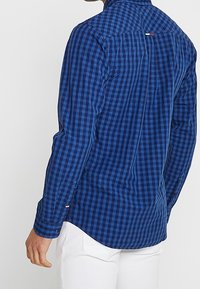Niebiesko-czarna koszula w kratę z długim rękawem, zaokrąglonym dołem, mankietami na guziki oraz metką z logo z tyłu przy kołnierzu. Tekstura bawełnianej tkaniny.