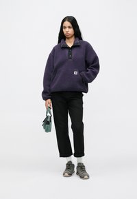 Naine seisab, seljas lilla fliisist sukka, mustad poolpüksid, hallid tossud, valged sokid ja hoiab käes väikest rohelist käekotti.