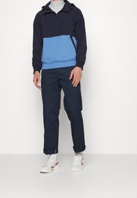 Marinblå halvdragkedja med huva, tillverkad av mjukt material; kombinerad med marinblå byxor och vita sneakers med röda detaljer.