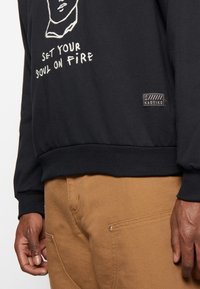 Sudadera negra con diseño blanco bordado que presenta una cara y el texto "ENCIENDE TU ALMA". Puños y dobladillo acanalados, y etiqueta de marca en el lateral.
