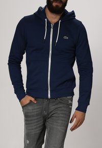 Marinblå zip-up hoodie tillverkad av mjukt material, med vita dragsko och en subtil logotyp på bröstet samt framfickor.