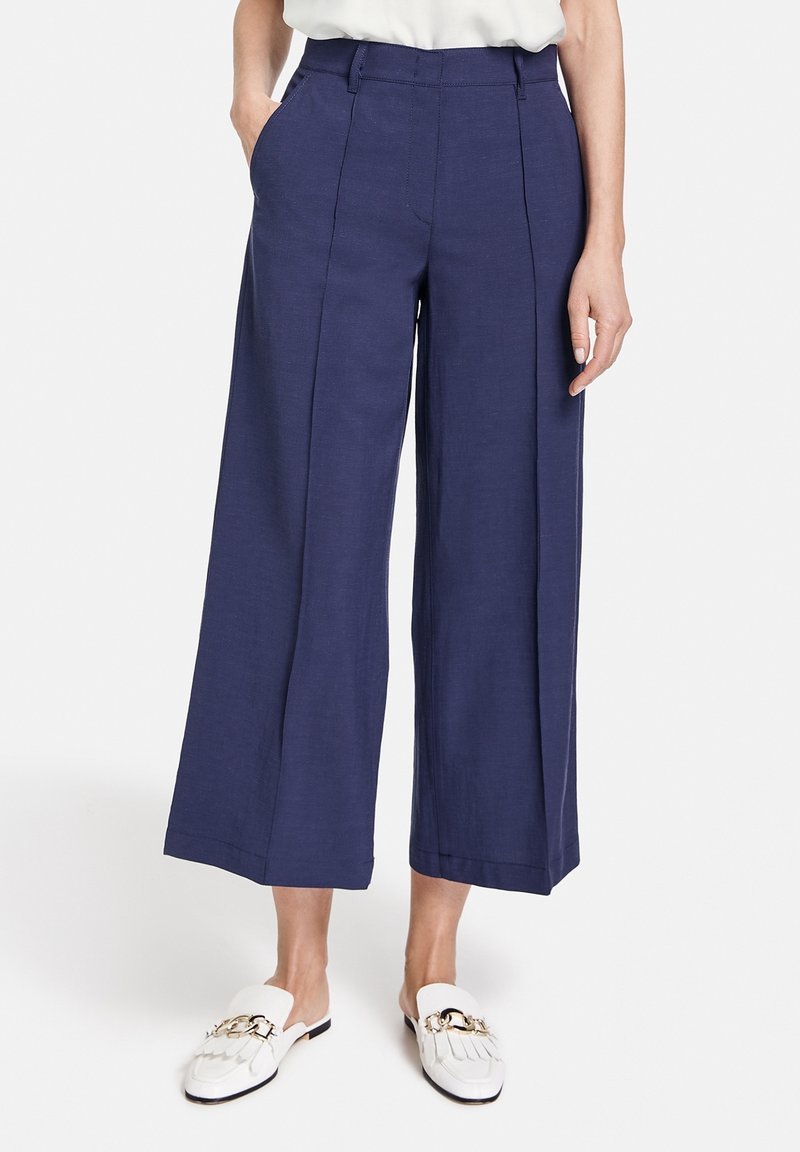 Marineblauwe wijde broek van een gladde stof met voorplooien en zijzakken, gecombineerd met witte instappers met gouden hardware accenten.