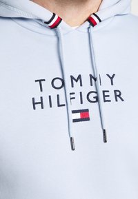 Sudadera azul clara con capucha, que presenta un gran logo bordado de "TOMMY HILFIGER" y un pequeño diseño de bandera cerca del dobladillo. Cordones con puntas metálicas.