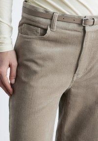 Beige Corduroyhose mit vertikalen Rippungen, einer Vorderseite-Tasche und einem passenden beigen Gürtel mit einer silbernen Schnalle. Weiche Textur sichtbar.