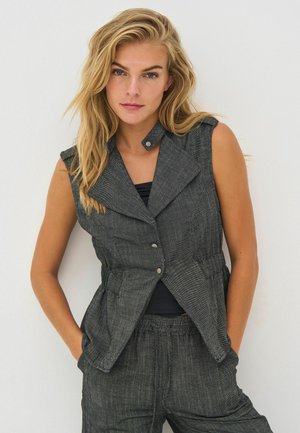 Szőke nő, laza hajjal, ujjatlan szürke mellényt és hozzá illő nadrágot visel, zsebre tett kézzel, fehér háttér előtt pózol.